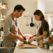 Un homme et une femme cuisinent ensemble dans une petite cuisine moderne en optimisant l’espace du plan de travail grâce à des rangements verticaux et des accessoires bien organisés.