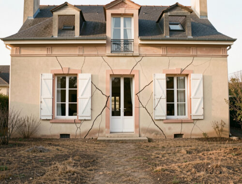 Façade d’une maison individuelle en France montrant des fissures obliques près des fenêtres, avec un jardin desséché après une période de sécheresse.